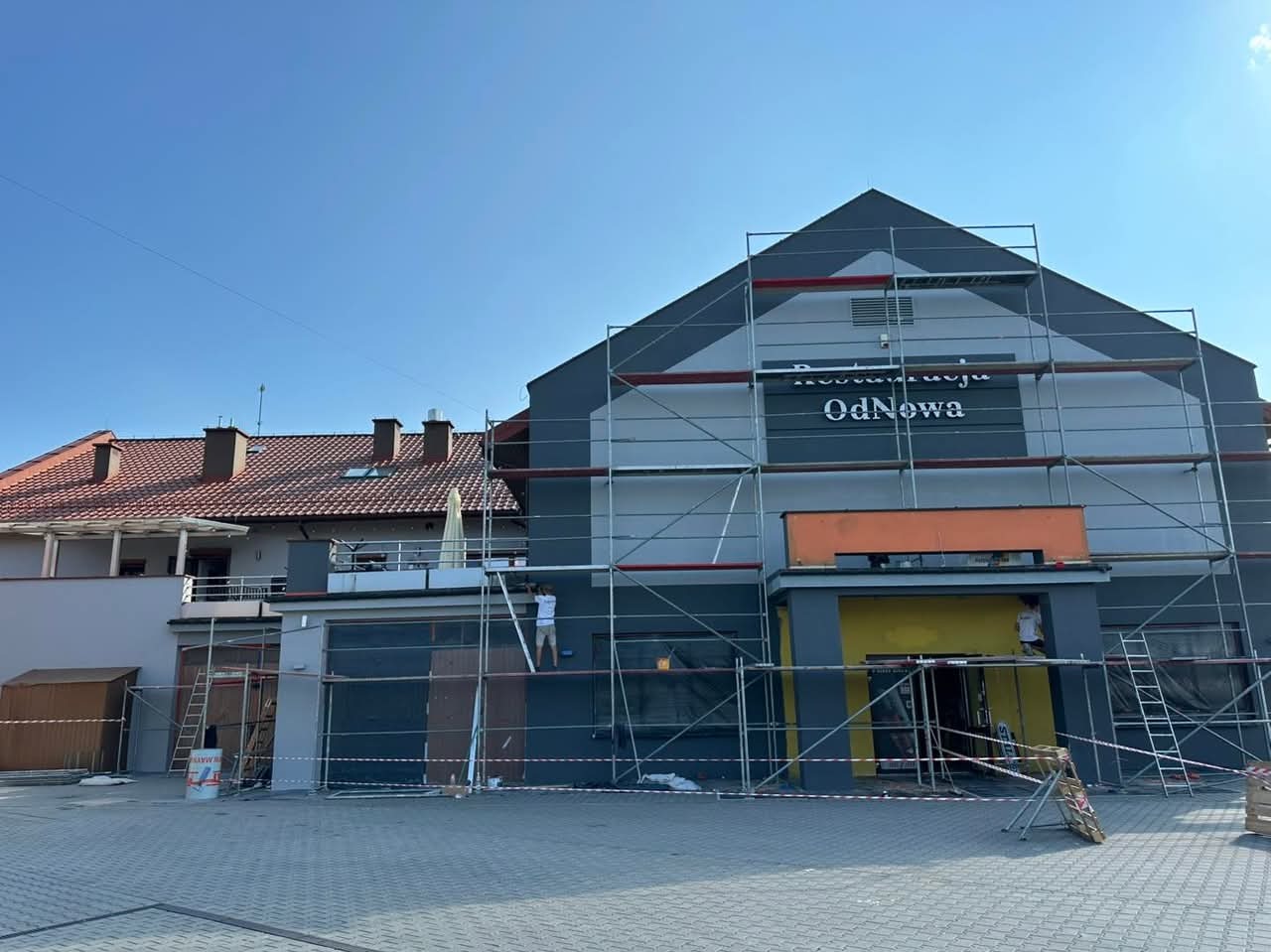 Dwukondygnacyjny budynek z napisem 'Restauracja OdNowa' na elewacji, otoczony rusztowaniami, dwóch pracowników w trakcie prac malarskich, widoczne puszki z farbą i drabina.