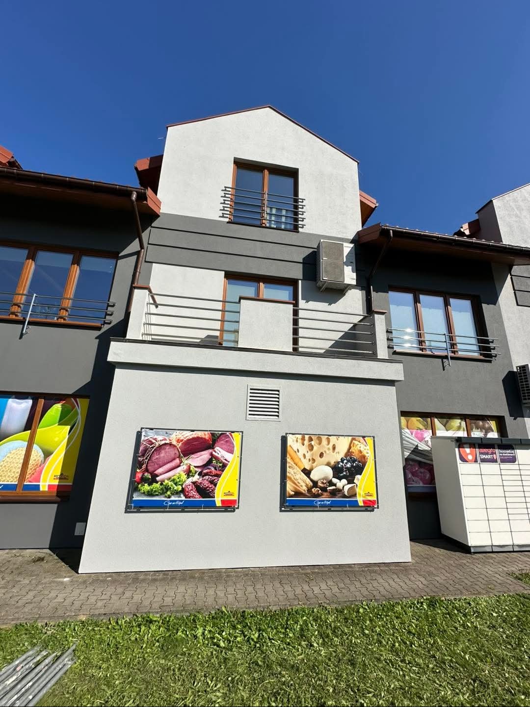 Dwukondygnacyjny budynek z szarą elewacją i dwoma reklamowymi banerami z jedzeniem na ścianie, widoczne okna z brązowymi ramami i balkon z metalową barierką.