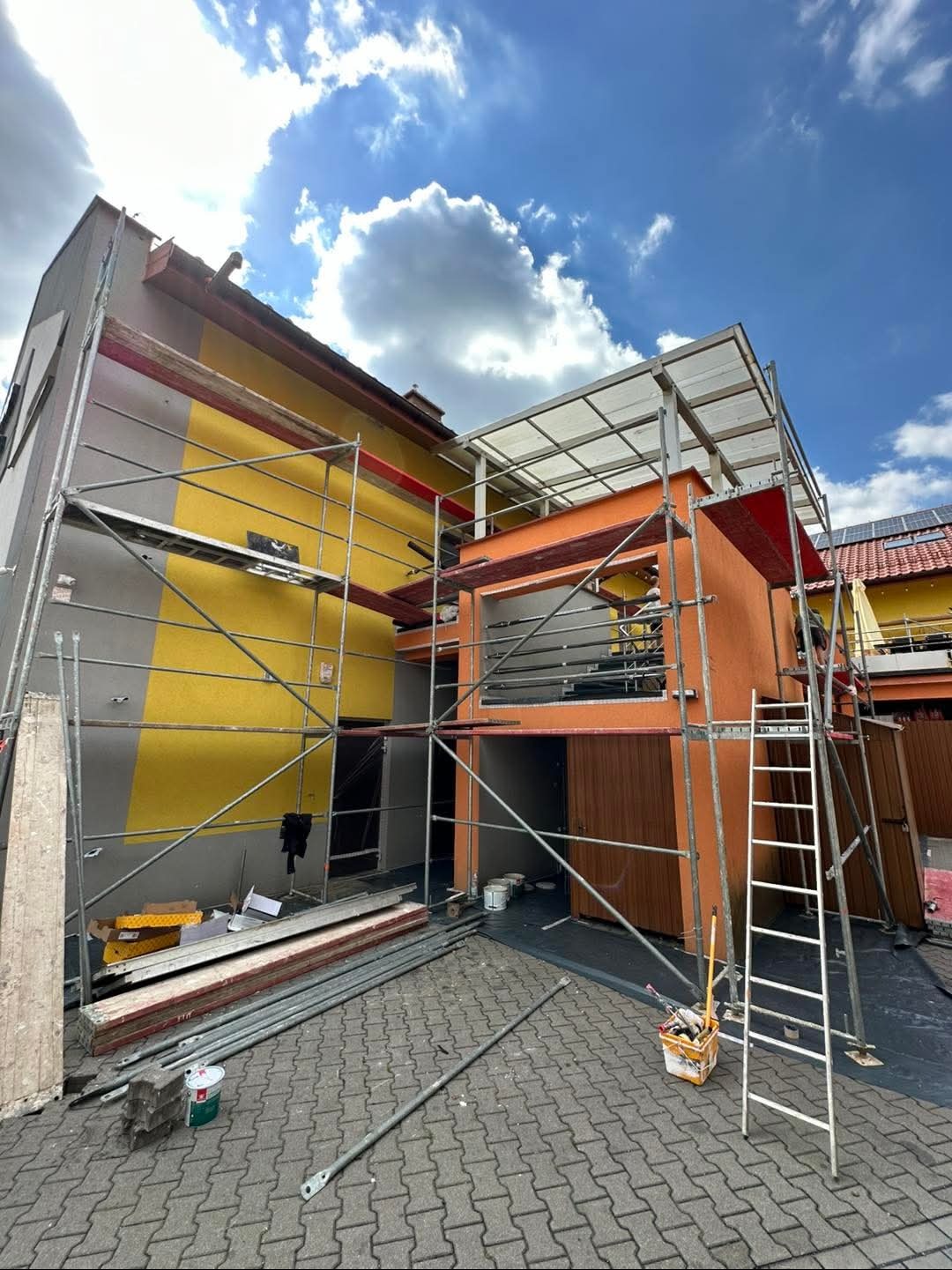 Budynek w trakcie prac elewacyjnych, z widocznym rusztowaniem, fragmentami otynkowanej i pomalowanej ściany w kolorach szarym i żółtym, oraz pomarańczową dobudówką z zadaszeniem. Na pierwszym...