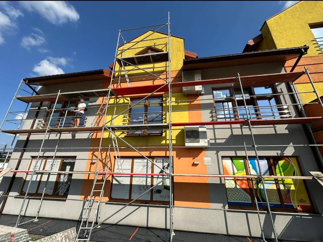 Budynek wielorodzinny w trakcie prac remontowych elewacji, widoczne rusztowania, fragmenty pomalowane na żółto, pomarańczowo i szaro, pracownik na rusztowaniu.