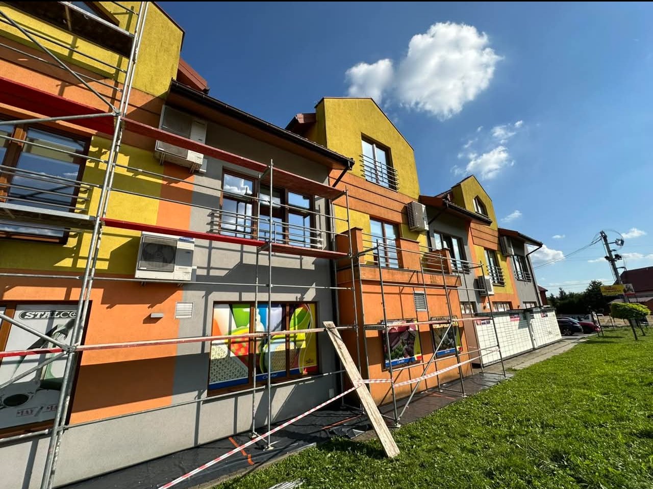 Wielobarwny budynek w trakcie prac elewacyjnych, widoczne rusztowania, klimatyzatory na ścianie i zabezpieczenia wokół budynku.