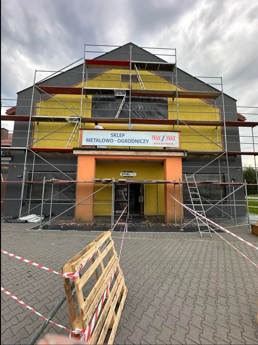 Sklep metalowo-ogrodniczy w trakcie prac elewacyjnych, widoczne rusztowania, częściowo pomalowana fasada na żółto i szaro, zabezpieczone okna folią, paleta na pierwszym planie.