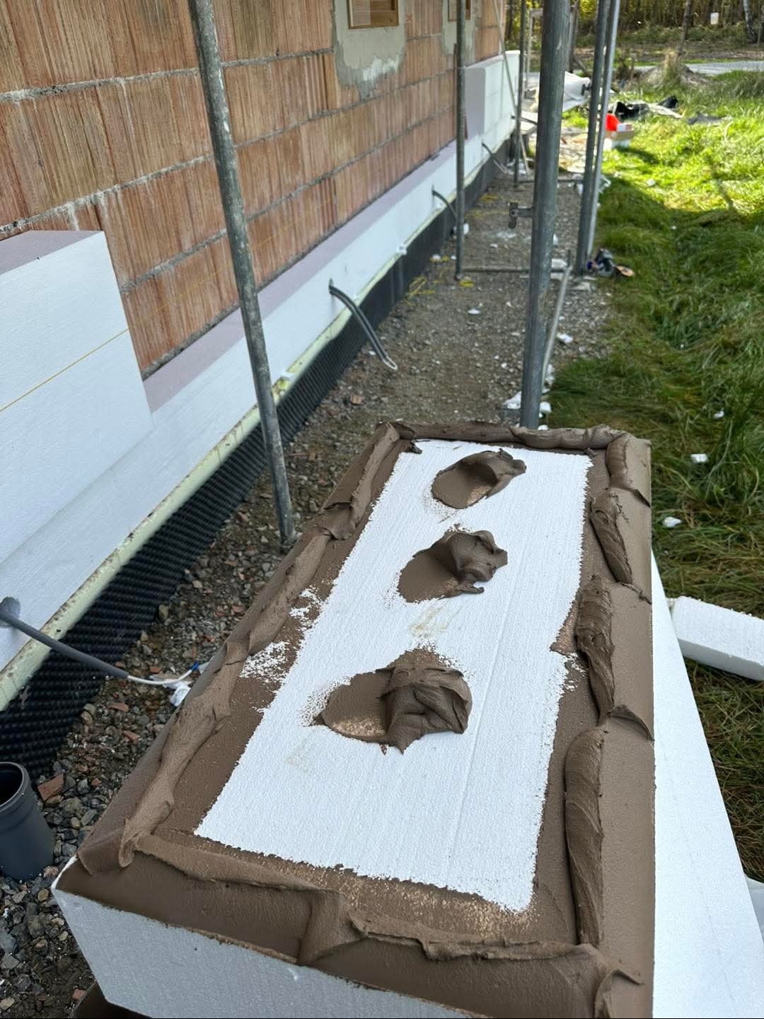 Przygotowanie styropianu do montażu na elewacji: biała płyta z trzema plamami brązowego kleju i obrzeżem z zaprawy, widoczna ściana budynku w trakcie ocieplania.