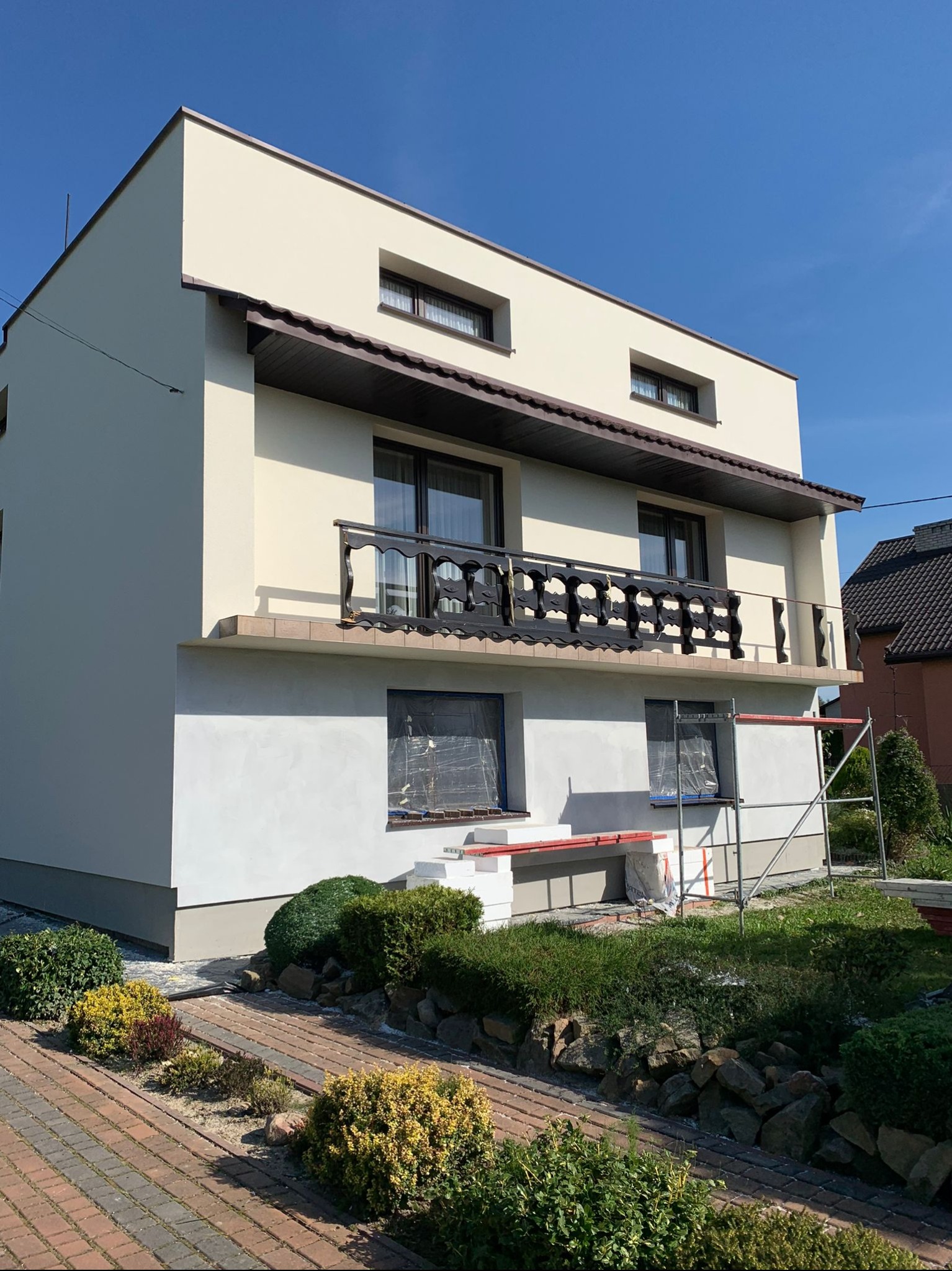 Dwukondygnacyjny dom jednorodzinny z balkonem w trakcie prac elewacyjnych, dolna część budynku w szarym tynku, górna w jasnym kremie, okna zabezpieczone folią, widoczne rusztowania i kostka brukowa...