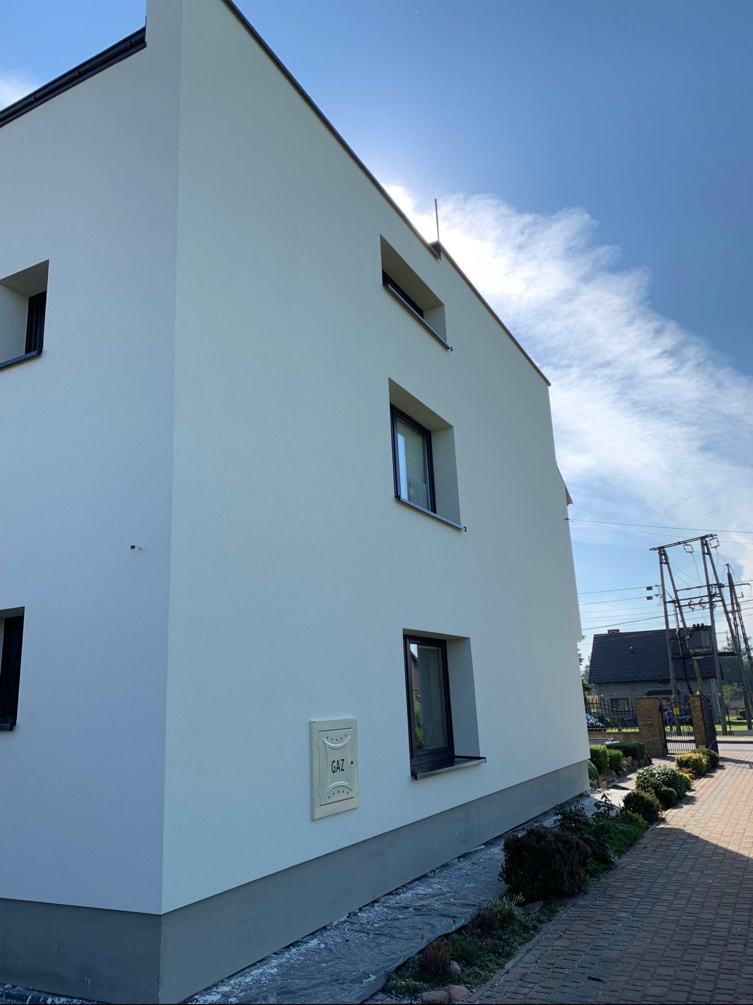 Nowoczesny, trzypiętrowy dom z białą elewacją i ciemnymi ramami okiennymi, widok z poziomu chodnika z kostki brukowej, obok skrzynka gazowa i skromna zieleń.