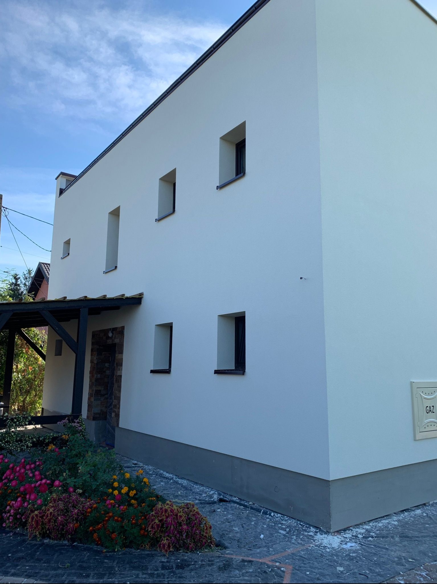 Biała, minimalistyczna elewacja domu jednorodzinnego z małymi oknami, widoczny fragment pergoli z kamiennym wykończeniem wejścia oraz rabata z kolorowymi kwiatami. Na ścianie skrzynka gazowa.