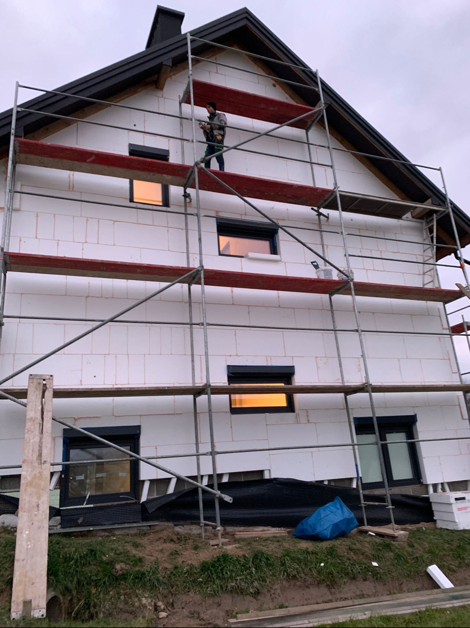 Pracownik na rusztowaniu, wykonuje prace na elewacji domu z białych bloczków, widoczne okna z roletami, oświetlone od wewnątrz, oraz zabezpieczenia gruntowe.