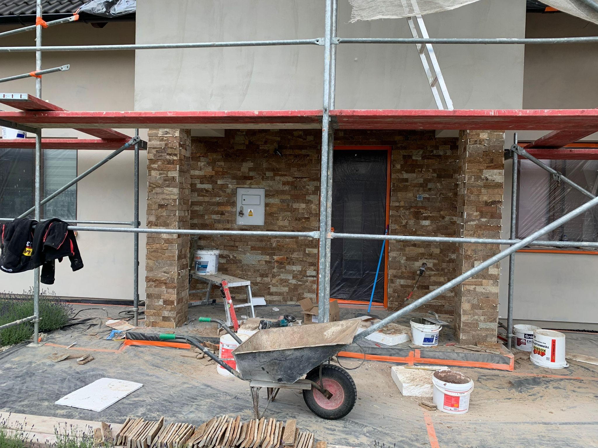 Montaż elewacji z kamienia dekoracyjnego na ścianie domu, widoczne rusztowanie, taczka, narzędzia i materiały budowlane, drzwi zabezpieczone folią.