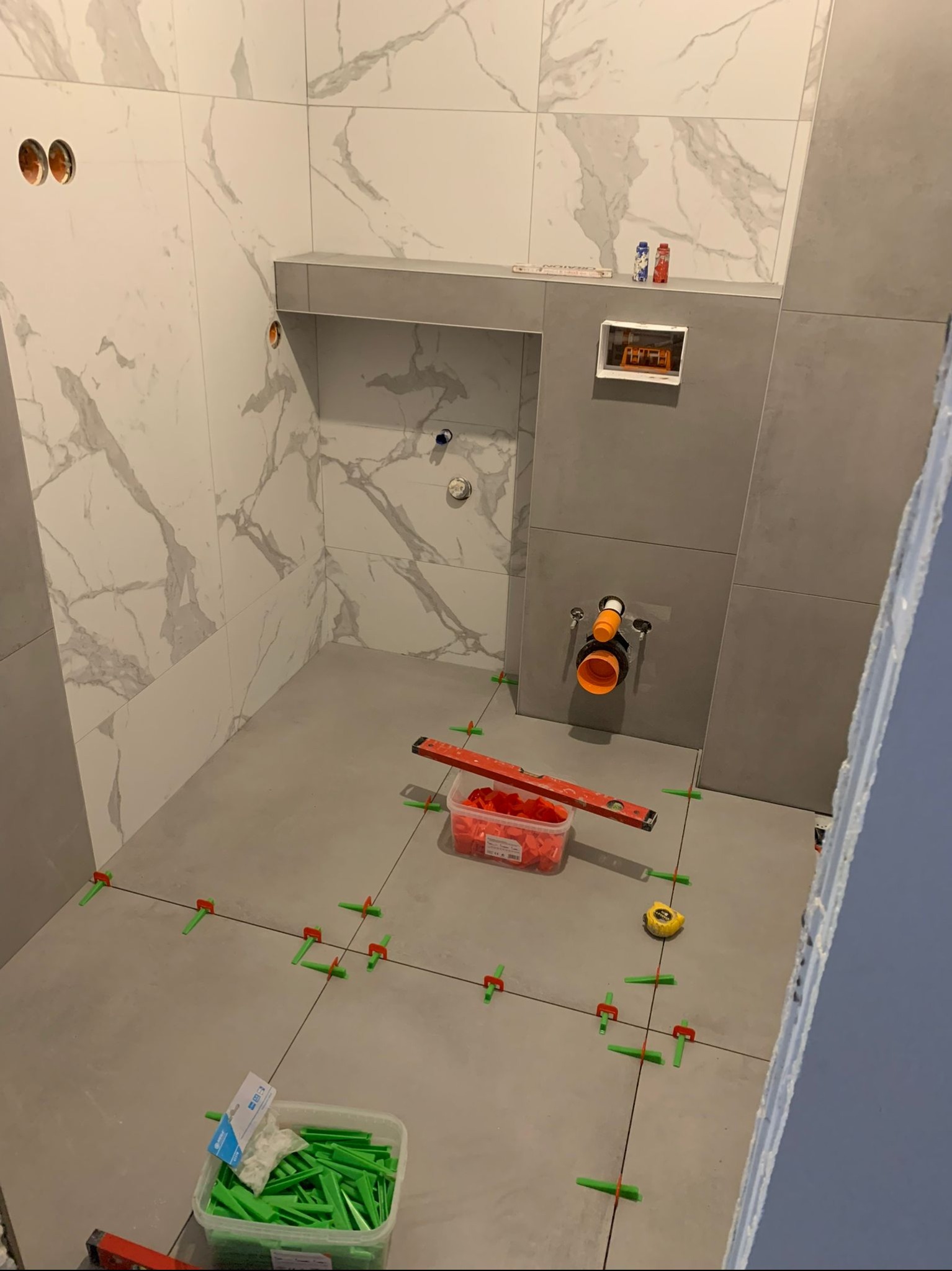 Łazienka w trakcie układania szarych płytek podłogowych i białych, marmurkowych płytek ściennych. Widoczne poziomica, miara, pojemniki z krzyżykami dystansowymi, rury i otwory instalacyjne.