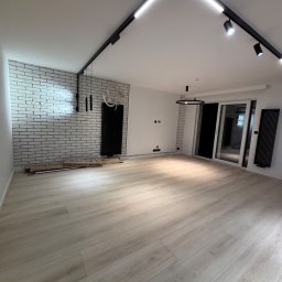 Wykończenia4you Sp. z o.o. - Wnętrze pokoju z wykończoną ścianą z białej cegły, panelami podłogowymi w odcieniach beżu i systemem oświetlenia szynowego na suficie.