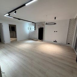 Wykończenia4you Sp. z o.o. - Jasne panele podłogowe w przestronnym pokoju z białymi ścianami, dekoracyjną ścianą z białej cegły, czarnym grzejnikiem i nowoczesnym oświetleniem sufitowym.