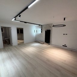 Wykończenia4you Sp. z o.o. - Jasne, przestronne wnętrze po remoncie z panelami podłogowymi, oświetleniem szynowym i dekoracyjną ścianą z białej cegły.