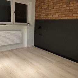 Wykończenia4you Sp. z o.o. - Jasne panele podłogowe w pokoju z ceglaną ścianą częściowo pomalowaną na czarno, biały grzejnik pod oknem.