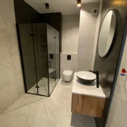 Nowoczesna łazienka z kabiną prysznicową typu walk-in z czarnymi profilami, umywalką nablatową w kształcie misy na szafce z drewnianym frontem i owalnym lustrem w czarnej ramie na ścianie...