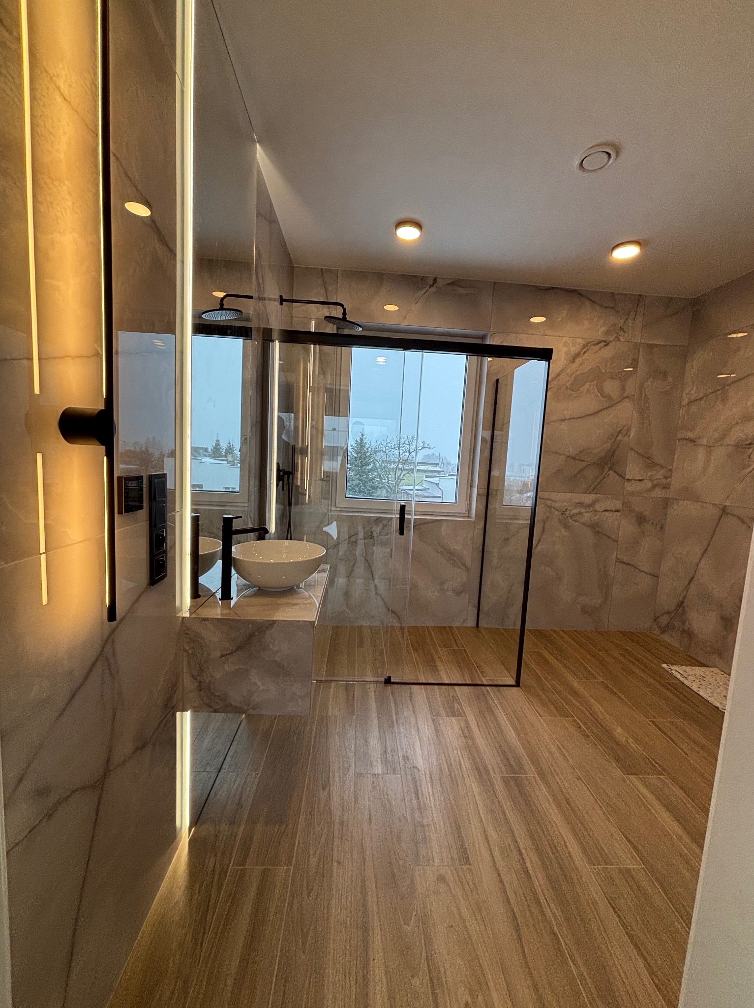 Nowoczesna łazienka z marmurowymi płytkami, drewnopodobną podłogą, kabiną prysznicową typu walk-in z czarnymi ramami i podwójną deszczownicą oraz umywalką nablatową na marmurowym blacie...