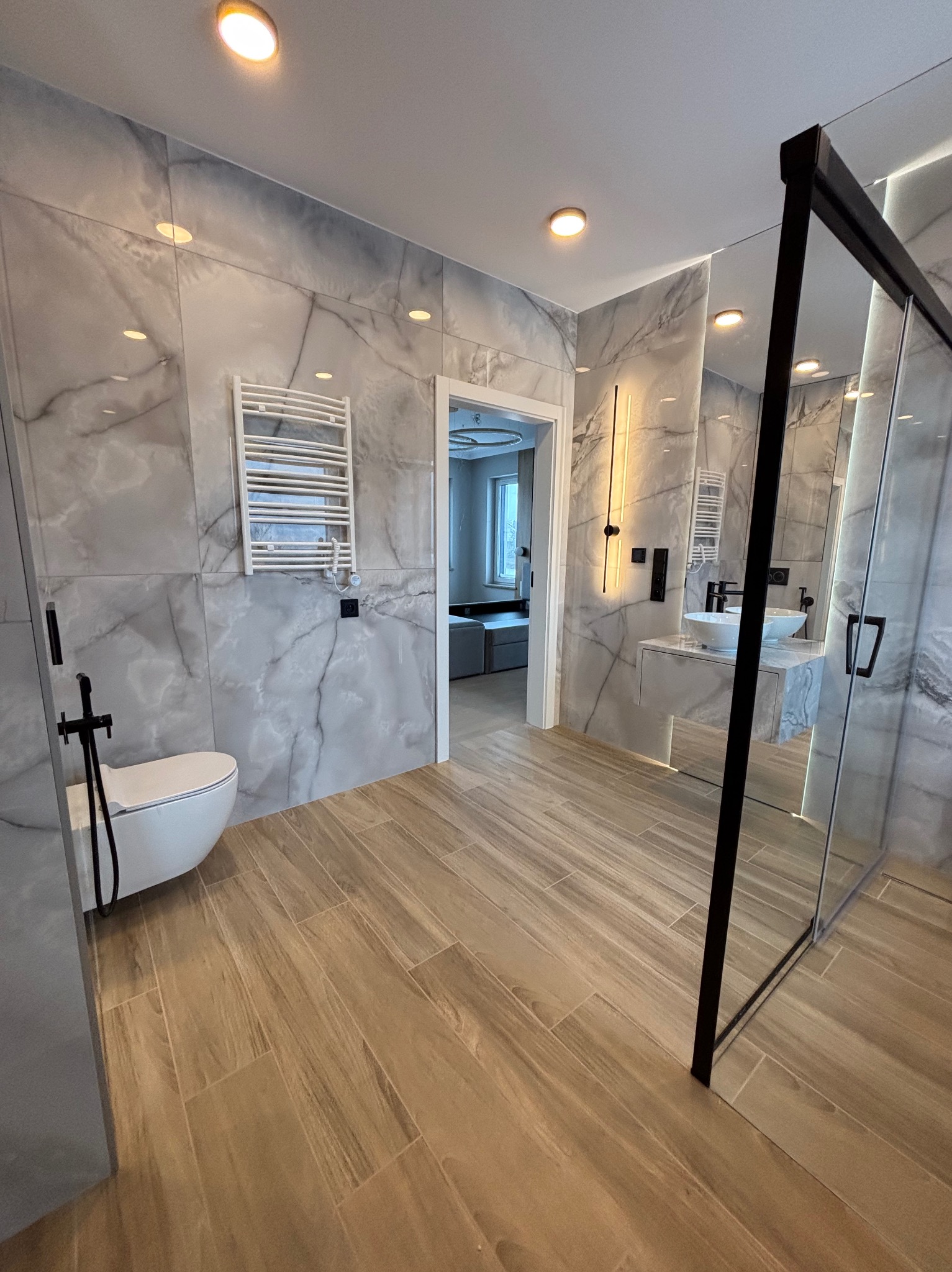 Nowoczesna łazienka z marmurowymi płytkami na ścianach, drewnopodobną podłogą, kabiną prysznicową z czarną ramą i minimalistyczną umywalką nablatową.