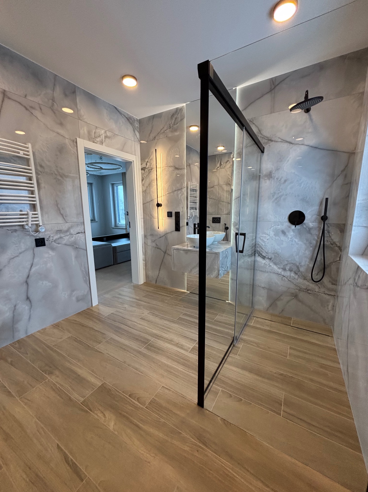 Nowoczesna łazienka z marmurowymi płytkami, kabiną prysznicową typu walk-in z czarną ramą, drewnopodobną podłogą i minimalistyczną armaturą, widok na pokój przez otwarte drzwi.