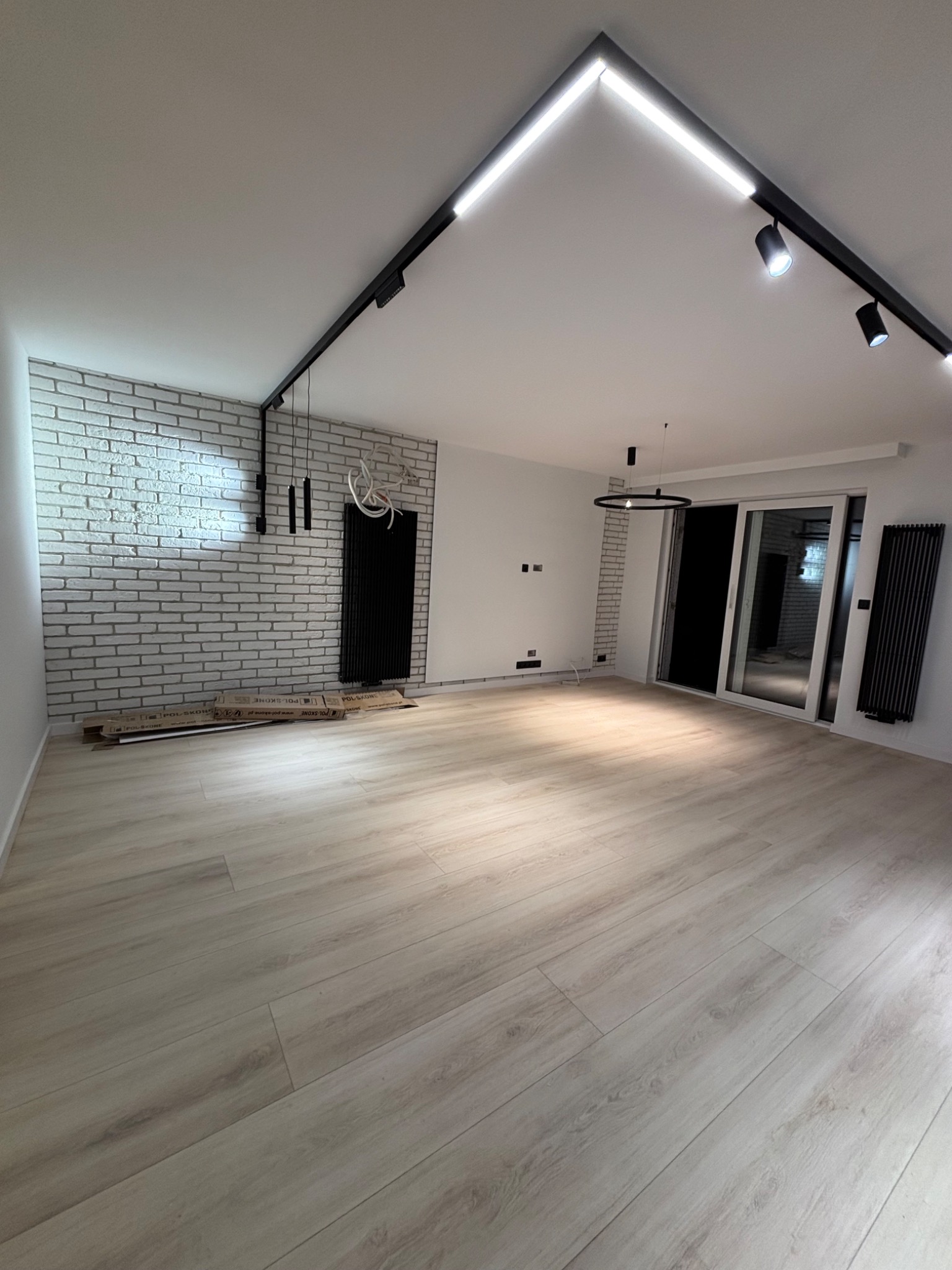 Wnętrze pokoju z wykończoną ścianą z białej cegły, panelami podłogowymi w odcieniach beżu i systemem oświetlenia szynowego na suficie.