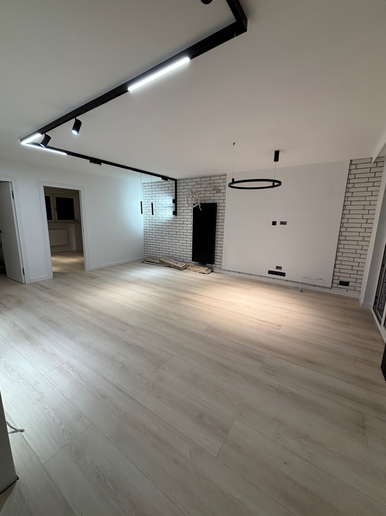 Jasne panele podłogowe w przestronnym pokoju z białymi ścianami, dekoracyjną ścianą z białej cegły, czarnym grzejnikiem i nowoczesnym oświetleniem sufitowym.