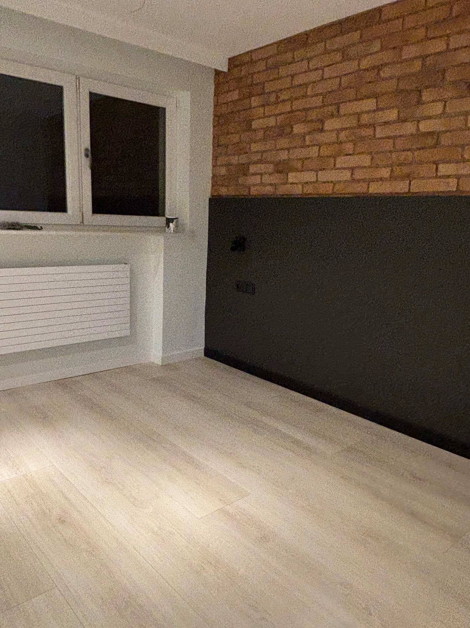 Jasne panele podłogowe w pokoju z ceglaną ścianą częściowo pomalowaną na czarno, biały grzejnik pod oknem.