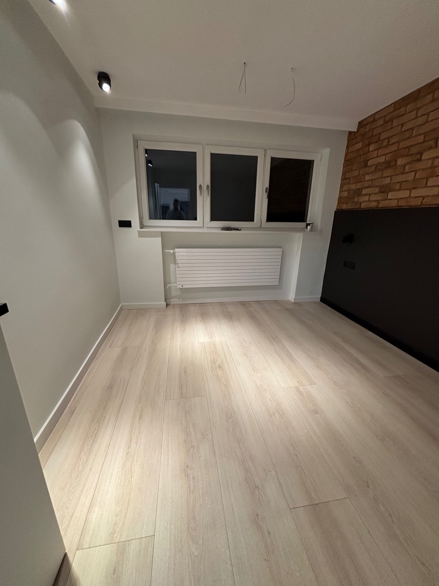 Puste pomieszczenie po remoncie z panelami podłogowymi w odcieniach beżu, białe ściany, grzejnik pod trzema oknami i fragment ściany z cegły z czarnym panelem dekoracyjnym.