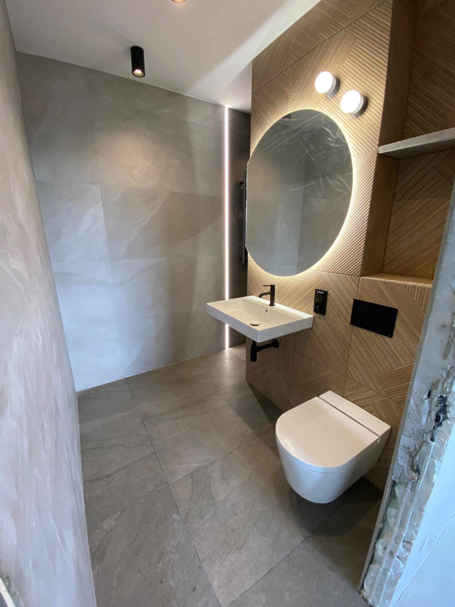 Nowoczesna łazienka z szarymi płytkami imitującymi kamień na podłodze i ścianach, drewnopodobną ścianą z geometrycznym wzorem, okrągłym lustrem z podświetleniem LED, wiszącą umywalką z czarną...