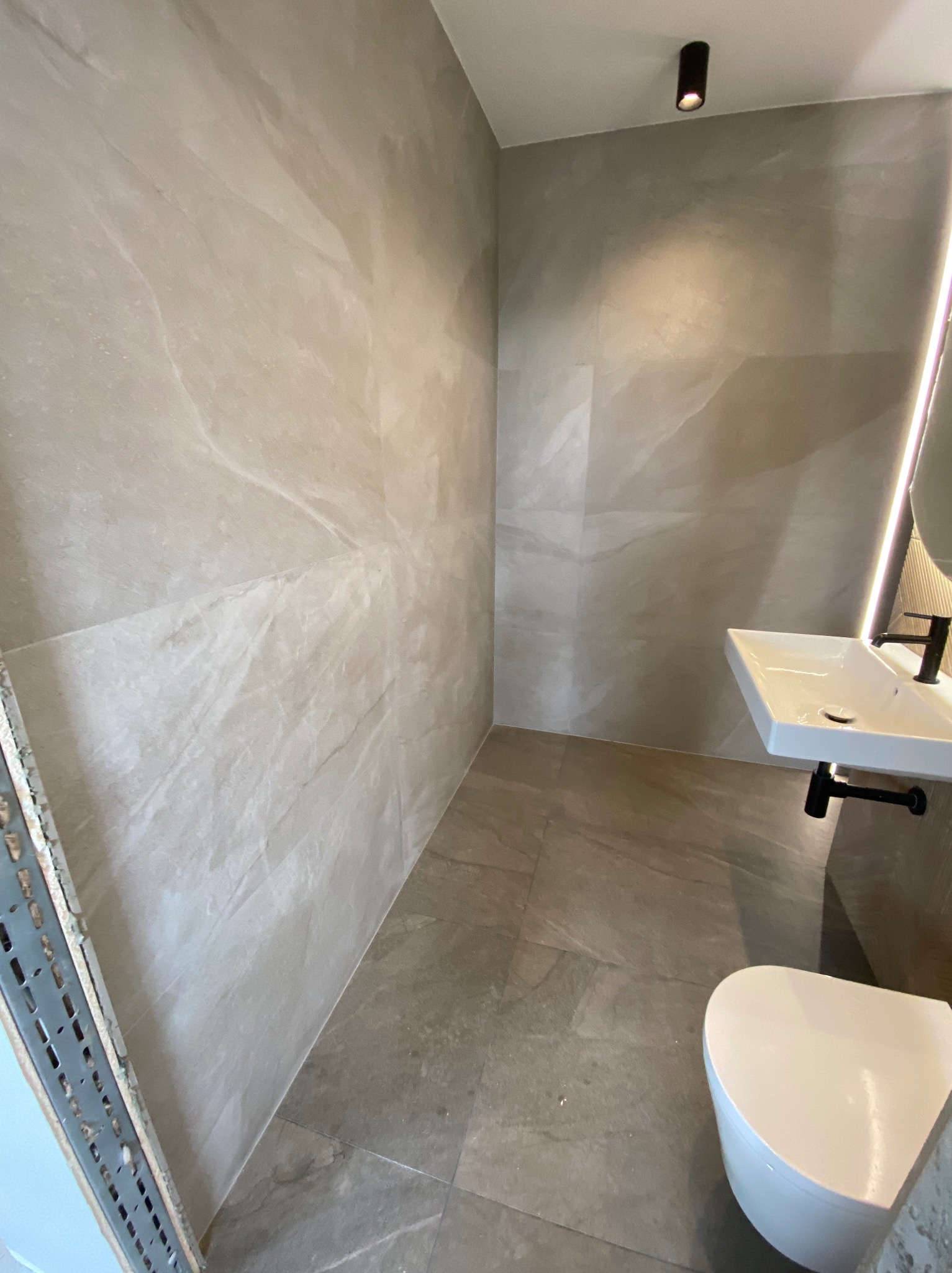 Nowoczesna łazienka w neutralnych odcieniach szarości i beżu, z podwieszaną umywalką, toaletą i dużymi płytami imitującymi marmur na ścianach i podłodze. Czarny punktowy reflektor sufitowy...