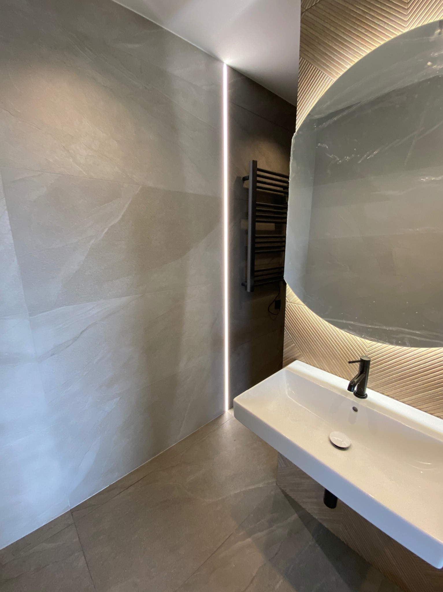 Nowoczesna łazienka z umywalką nablatową, podświetlanym lustrem w folii ochronnej i grzejnikiem drabinkowym na ścianie z płytek w odcieniach szarości i beżu.