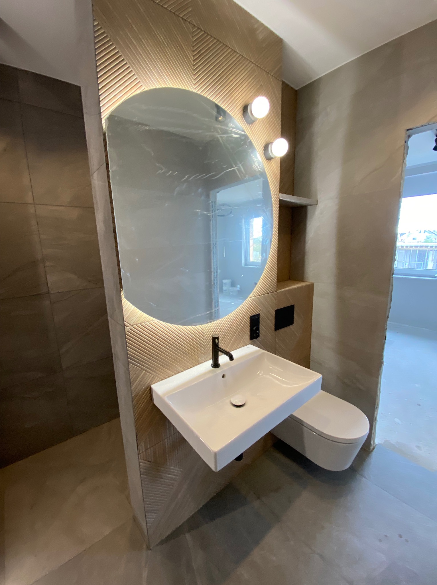 Nowoczesna łazienka z owalnym lustrem podświetlanym LED i wiszącą umywalką z czarną baterią, płytki imitujące drewno na ścianie i podłodze, toaleta podwieszana w tle.