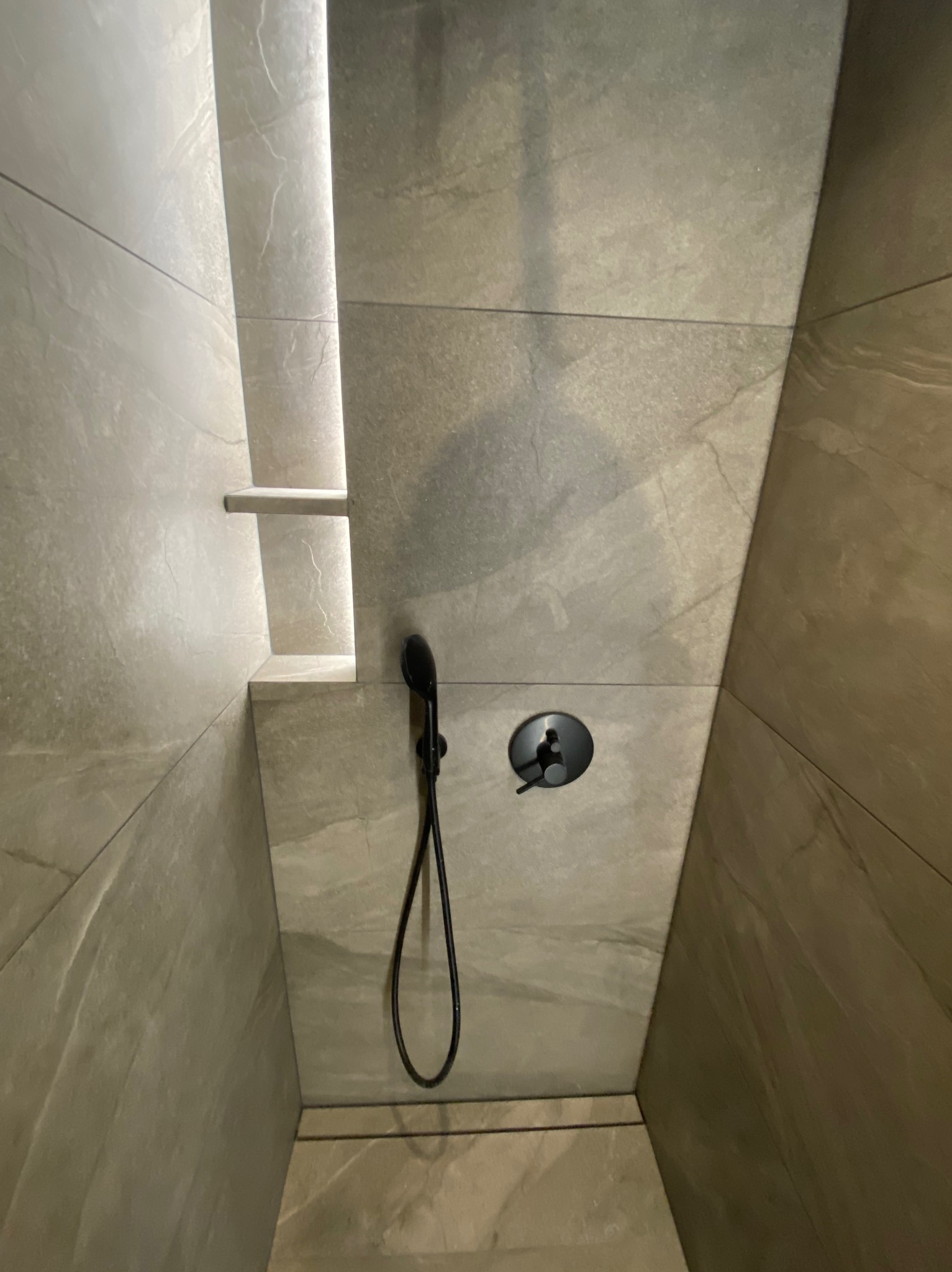 Nowoczesny prysznic z szarymi płytkami imitującymi kamień, czarną armaturą i podświetlaną półką wnękową.