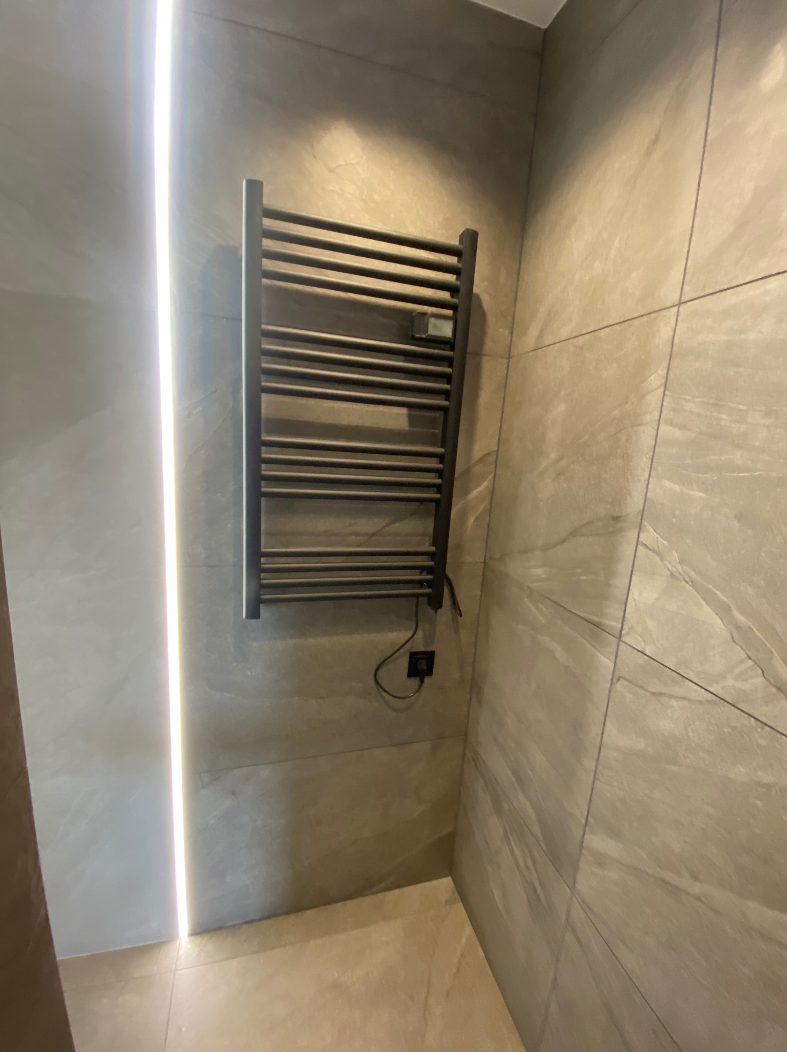 Nowoczesny, czarny grzejnik drabinkowy na ścianie z płytek imitujących kamień, podświetlonej pionową listwą LED w łazience.