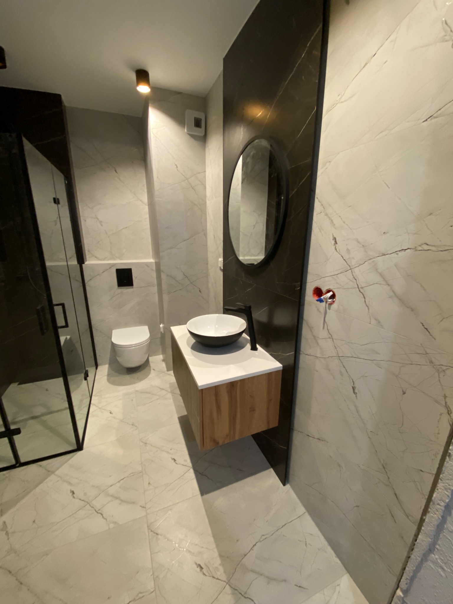 Nowoczesna łazienka z marmurowymi płytkami, wiszącą szafką z umywalką w kolorze drewna, okrągłym lustrem w czarnej ramie i czarną kabiną prysznicową walk-in.