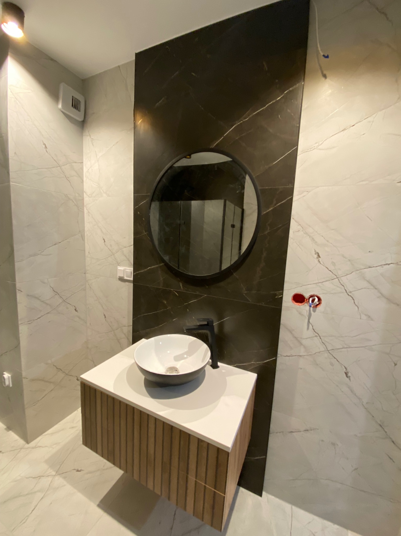 Nowoczesna łazienka z umywalką nablatową, okrągłym lustrem i marmurowymi płytkami w odcieniach bieli i czerni, widoczne przygotowane punkty podłączenia armatury.