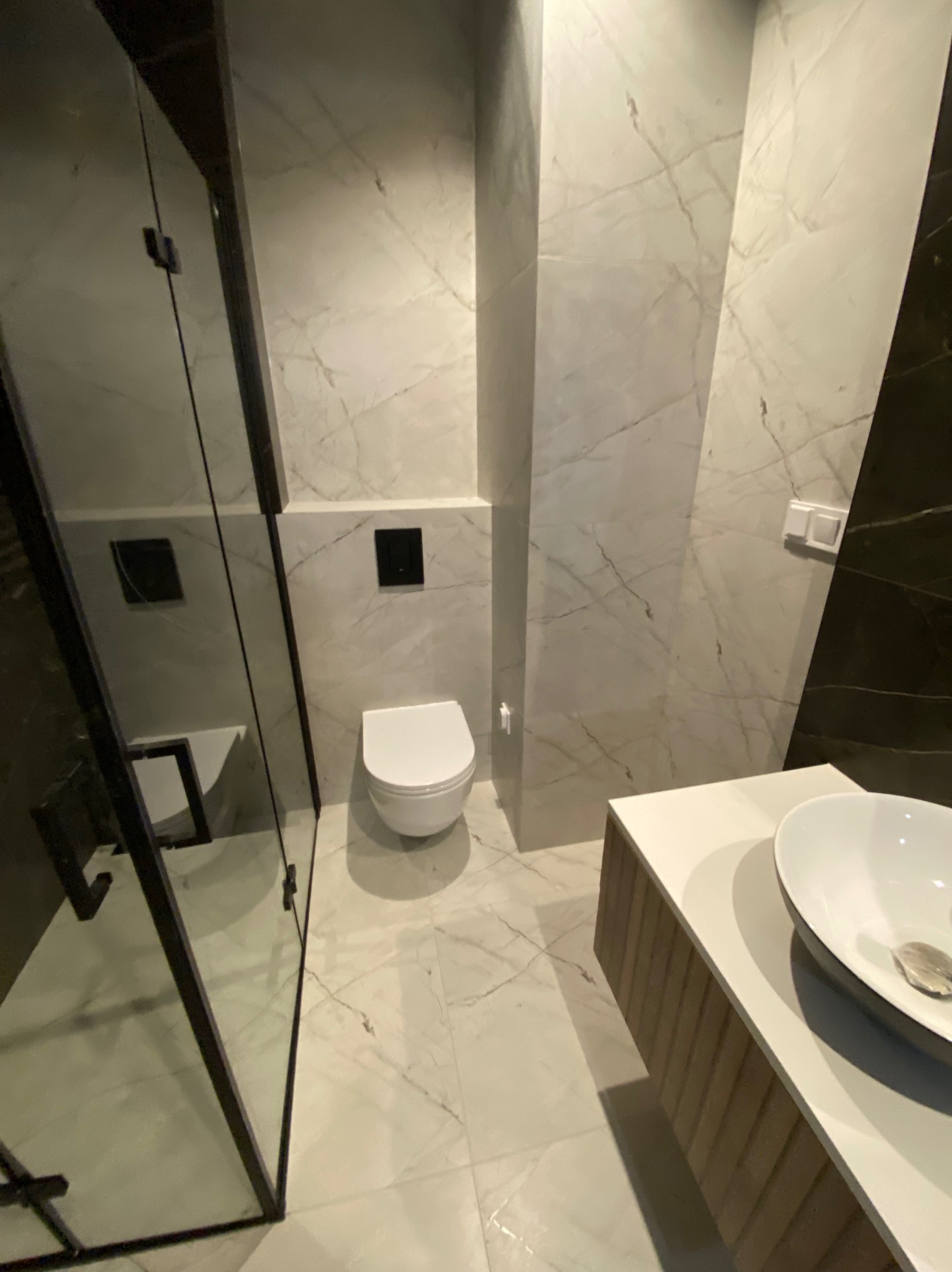 Nowoczesna łazienka z marmurowymi płytkami, kabiną prysznicową ze szkła hartowanego z czarnymi ramami, podwieszaną toaletą i umywalką nablatową z szafką z lamelami.