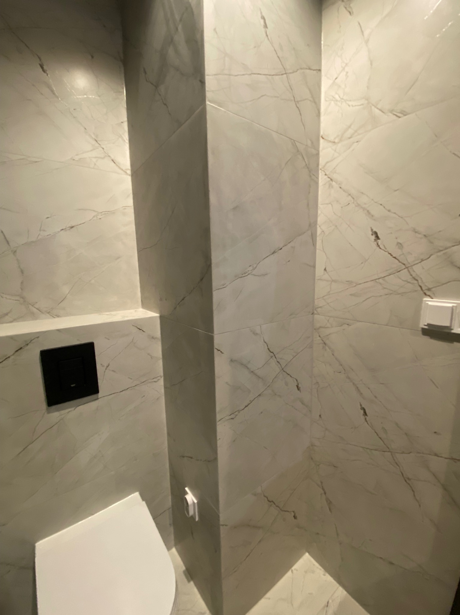 Wykończona łazienka z marmurowymi płytkami, podwieszaną toaletą z czarnym przyciskiem spłukiwania i białym, prostokątnym włącznikiem światła.