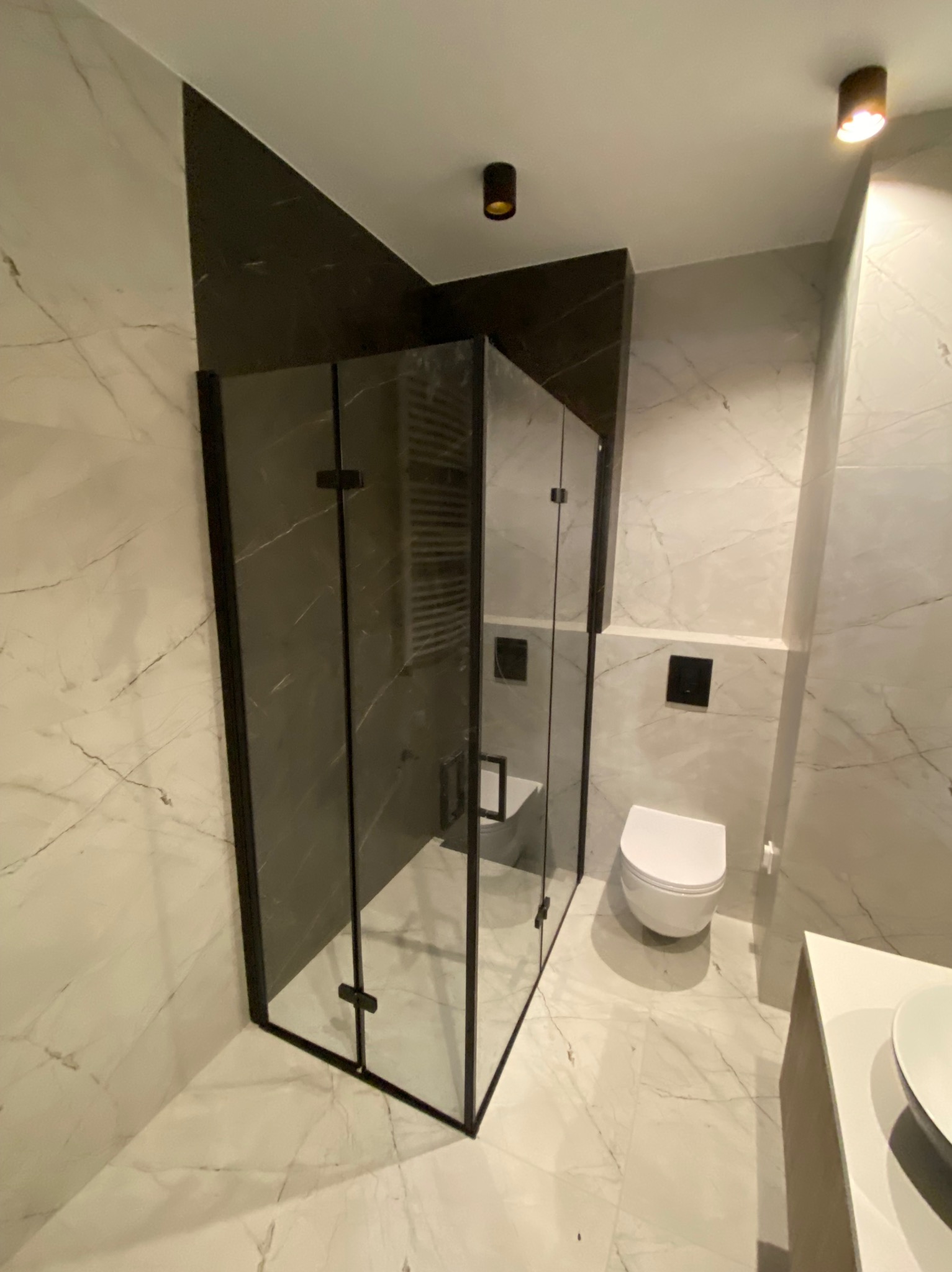 Nowoczesna łazienka z kabiną prysznicową z czarnymi ramami, marmurowymi płytkami i podwieszaną toaletą.