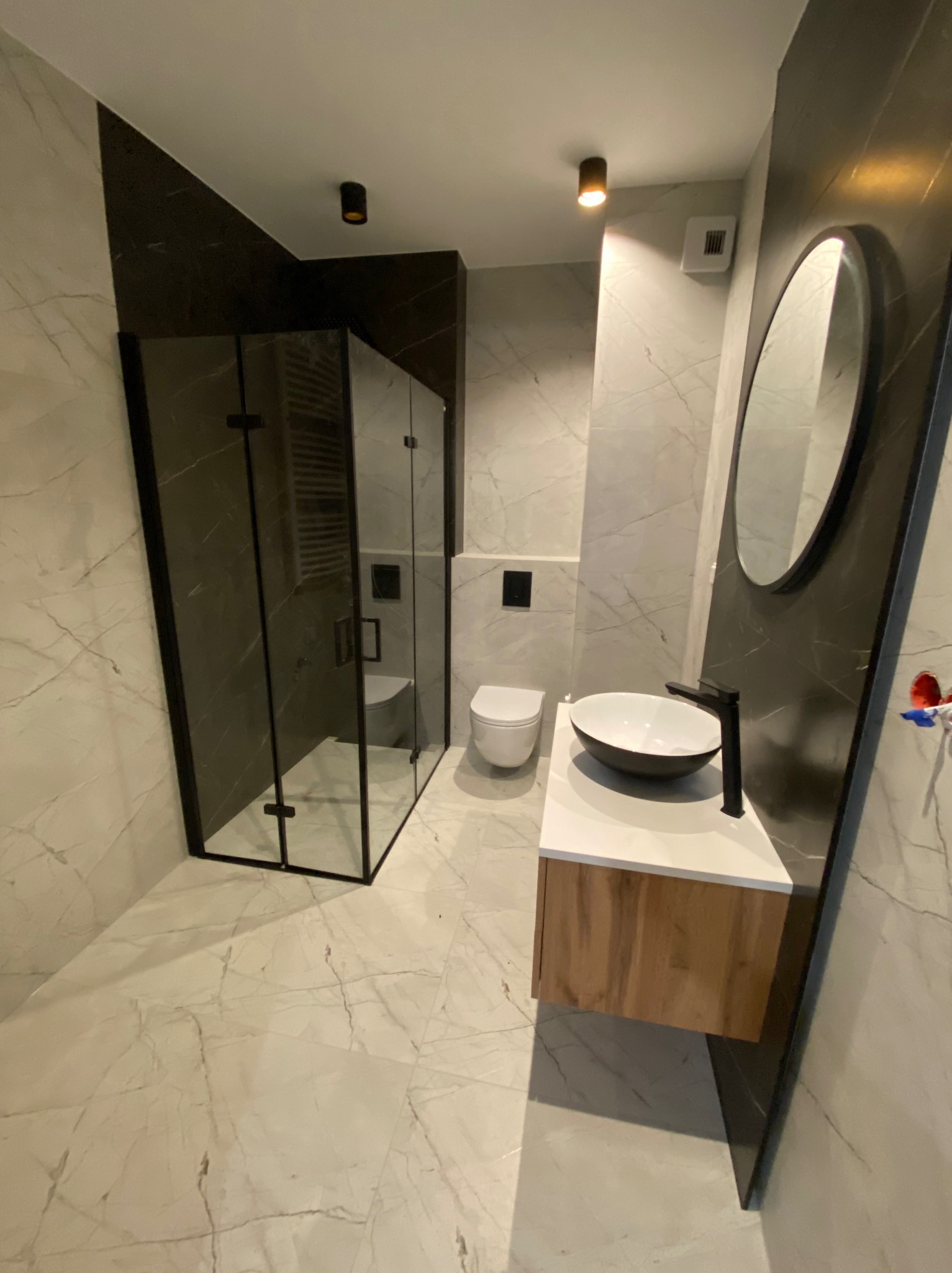 Nowoczesna łazienka z kabiną prysznicową typu walk-in z czarnymi profilami, umywalką nablatową w kształcie misy na szafce z drewnianym frontem i owalnym lustrem w czarnej ramie na ścianie...