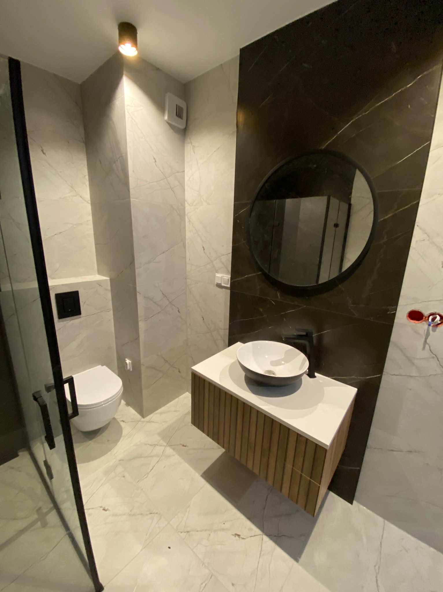 Nowoczesna łazienka z marmurowymi płytkami, wiszącą toaletą, umywalką nablatową na szafce z drewnianymi lamelami i okrągłym lustrem na ciemnej ścianie.