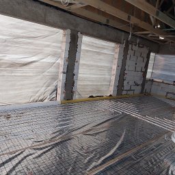 Andrii Shevchenko - Montaż ogrzewania podłogowego w surowym wnętrzu: widoczne rury na siatce, ściany z pustaków, folia ochronna na oknach i zwój rur obok kartonowych pudeł.