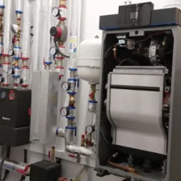 Nowoczesna kotłownia z piecem gazowym, rozdzielaczami Buderus, orurowaniem w izolacji i żółtą rurą gazową z oznaczeniem, widok całości instalacji.