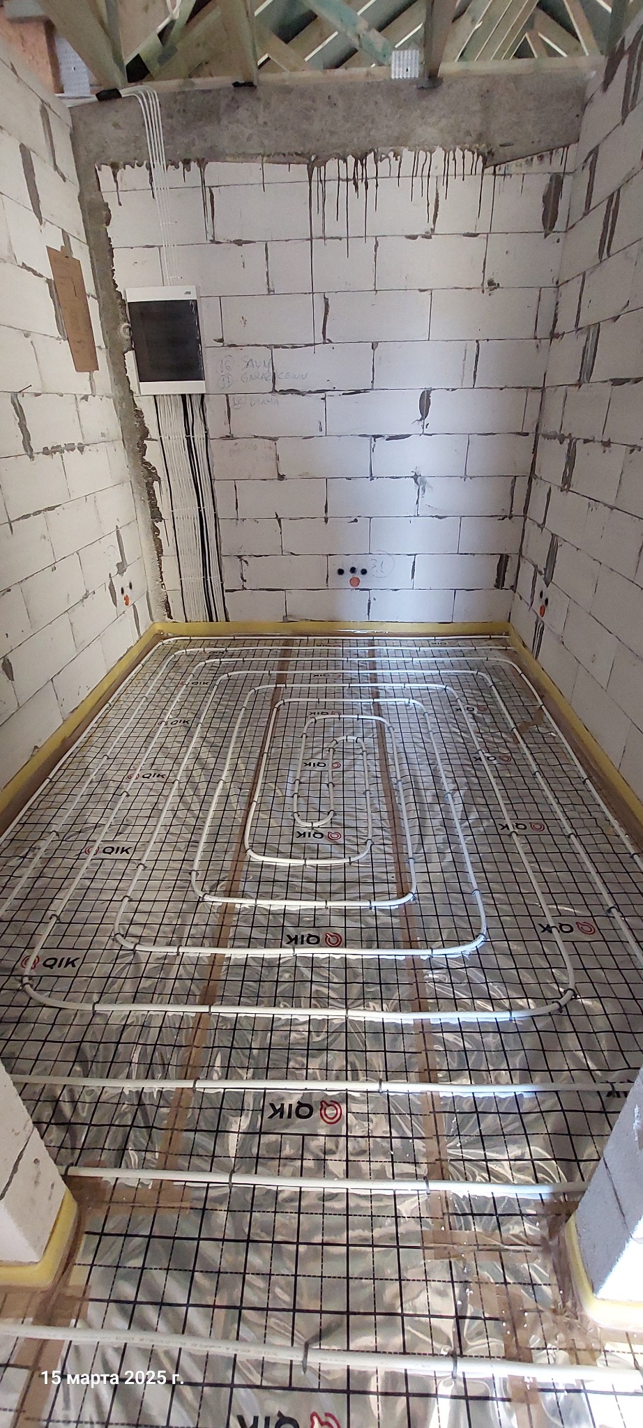 Białe rury ogrzewania podłogowego ułożone na srebrnej folii z czarną siatką w surowym pomieszczeniu z białych pustaków, widoczne przewody elektryczne i drewniana konstrukcja dachu.