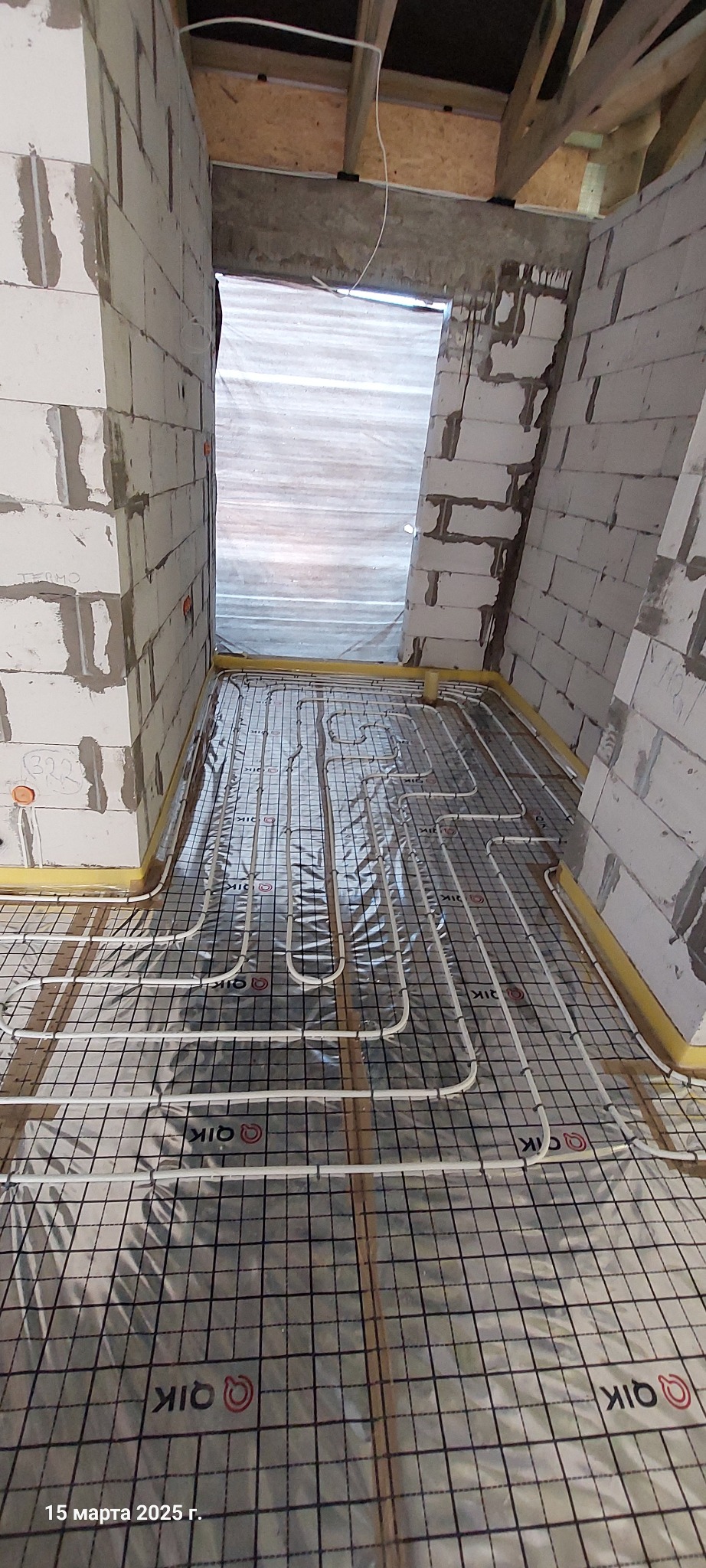 Układanie rur ogrzewania podłogowego na folii izolacyjnej z siatką, widoczne białe ściany z bloczków i surowy strop.