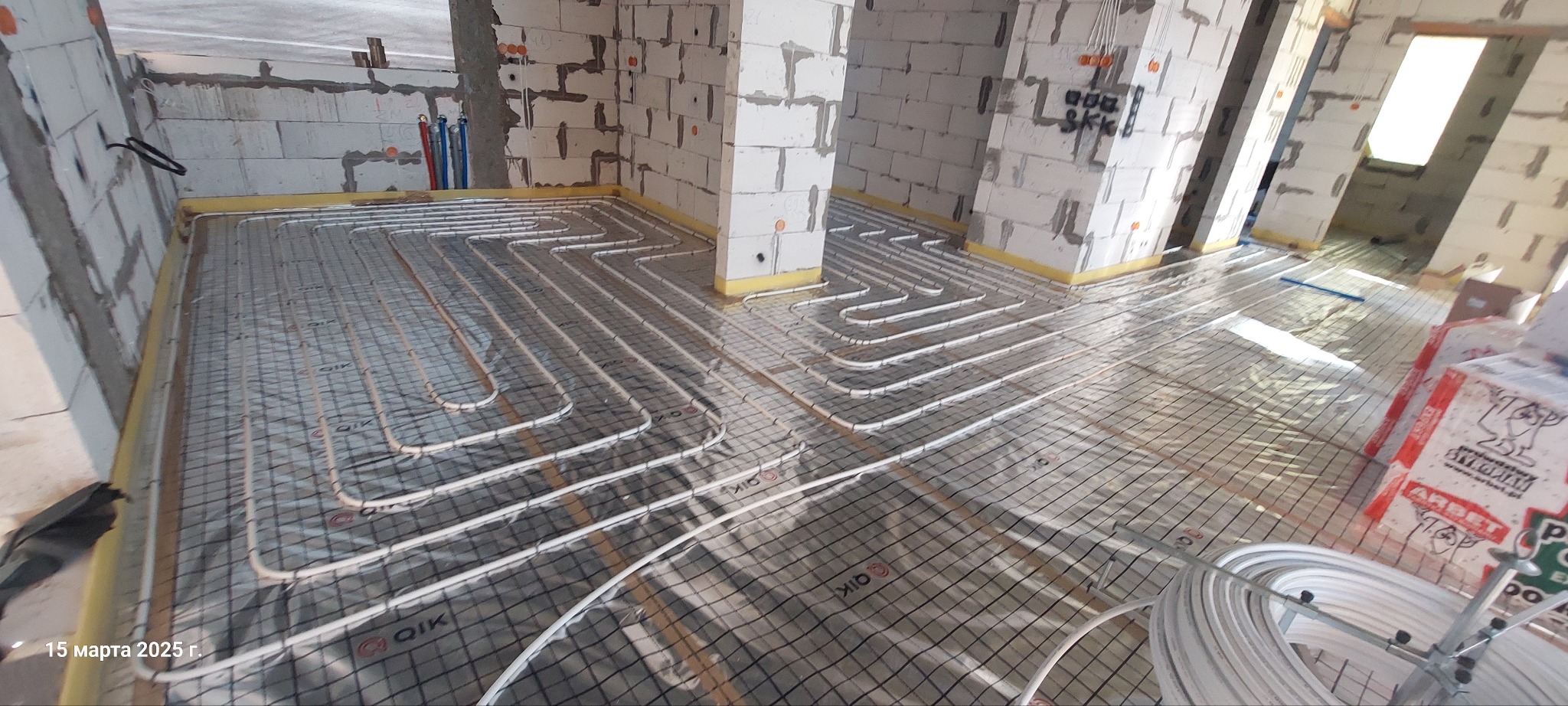 Białe rurki ogrzewania podłogowego ułożone na siatce w surowym wnętrzu budynku z białych bloczków, widoczne materiały budowlane i instalacje.