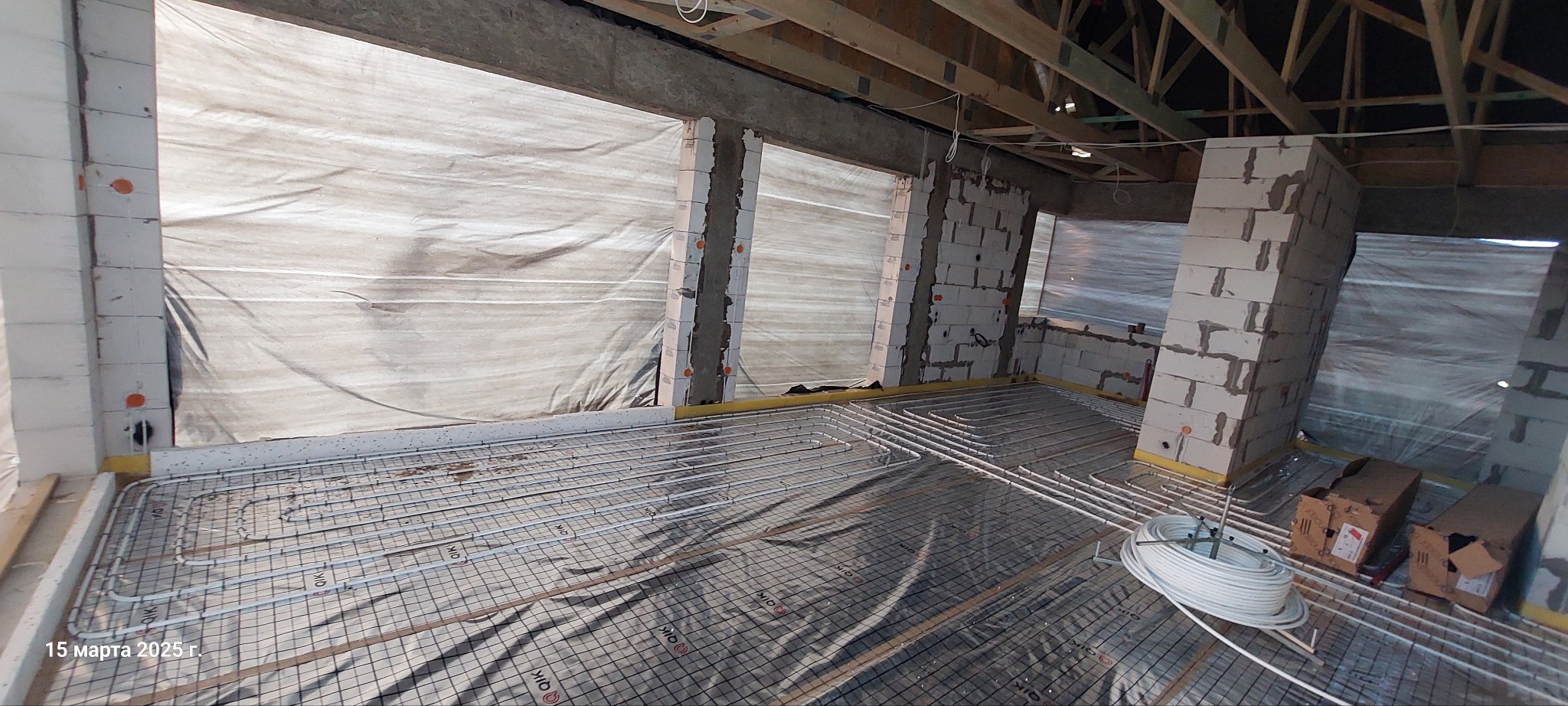 Montaż ogrzewania podłogowego w surowym wnętrzu: widoczne rury na siatce, ściany z pustaków, folia ochronna na oknach i zwój rur obok kartonowych pudeł.