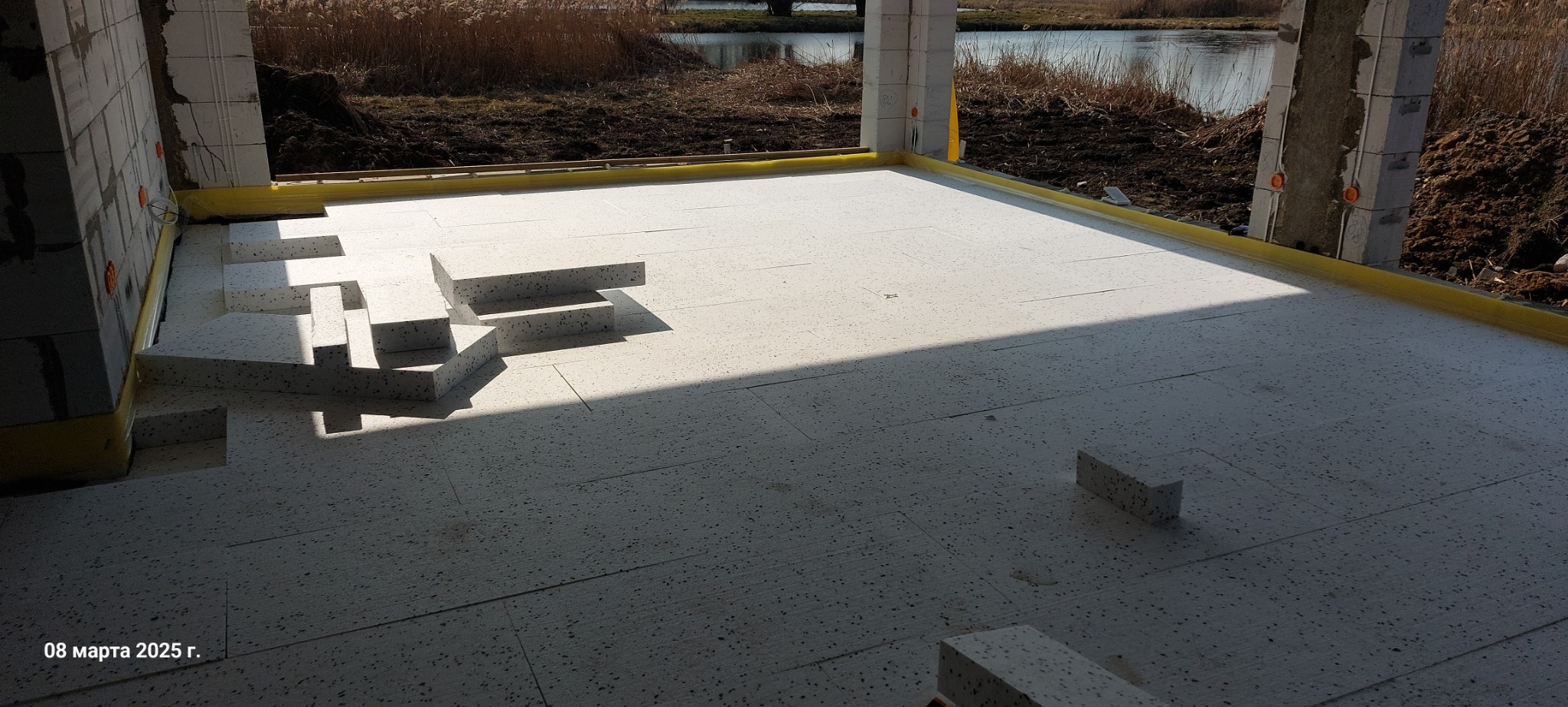 Montaż izolacji podłogowej z płyt styropianowych na przygotowanym podłożu w surowym stanie budynku, widoczne fragmenty ścian i słupów, na zewnątrz widok na jezioro.