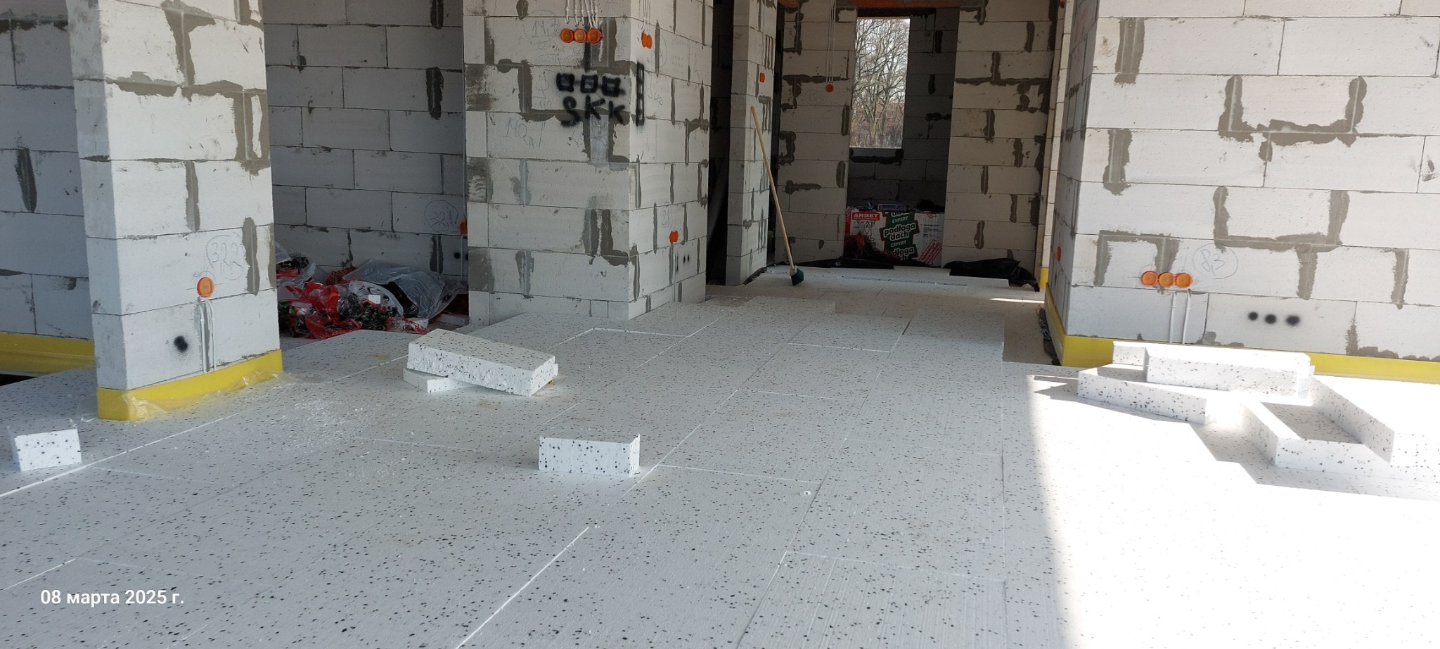 Wnętrze surowego domu w trakcie prac instalacyjnych, widoczne płyty styropianowe ułożone na podłodze, ściany z betonu komórkowego z widocznymi puszkami instalacji elektrycznej, żółta folia...