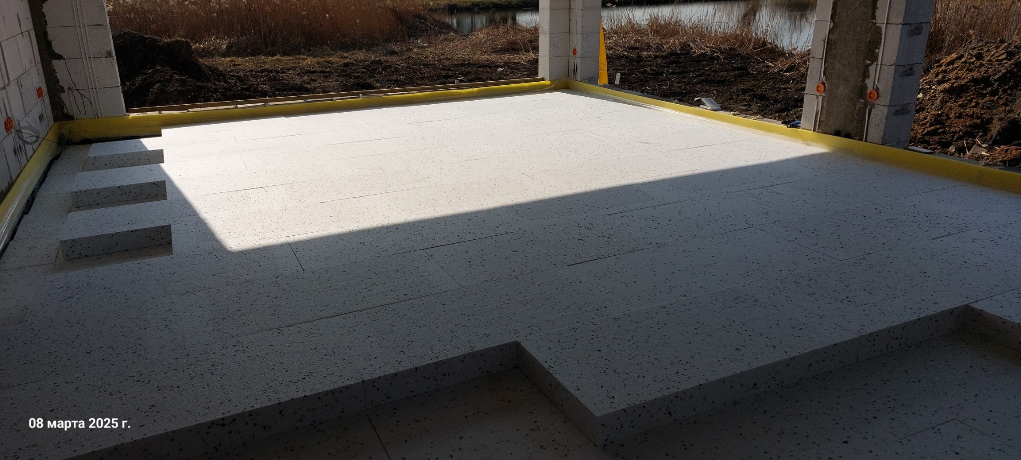 Izolacja podłogowa ze styropianu ułożona na przygotowanym podłożu w surowym stanie, widoczne żółte taśmy izolacyjne przy ścianach oraz wystające kable elektryczne.