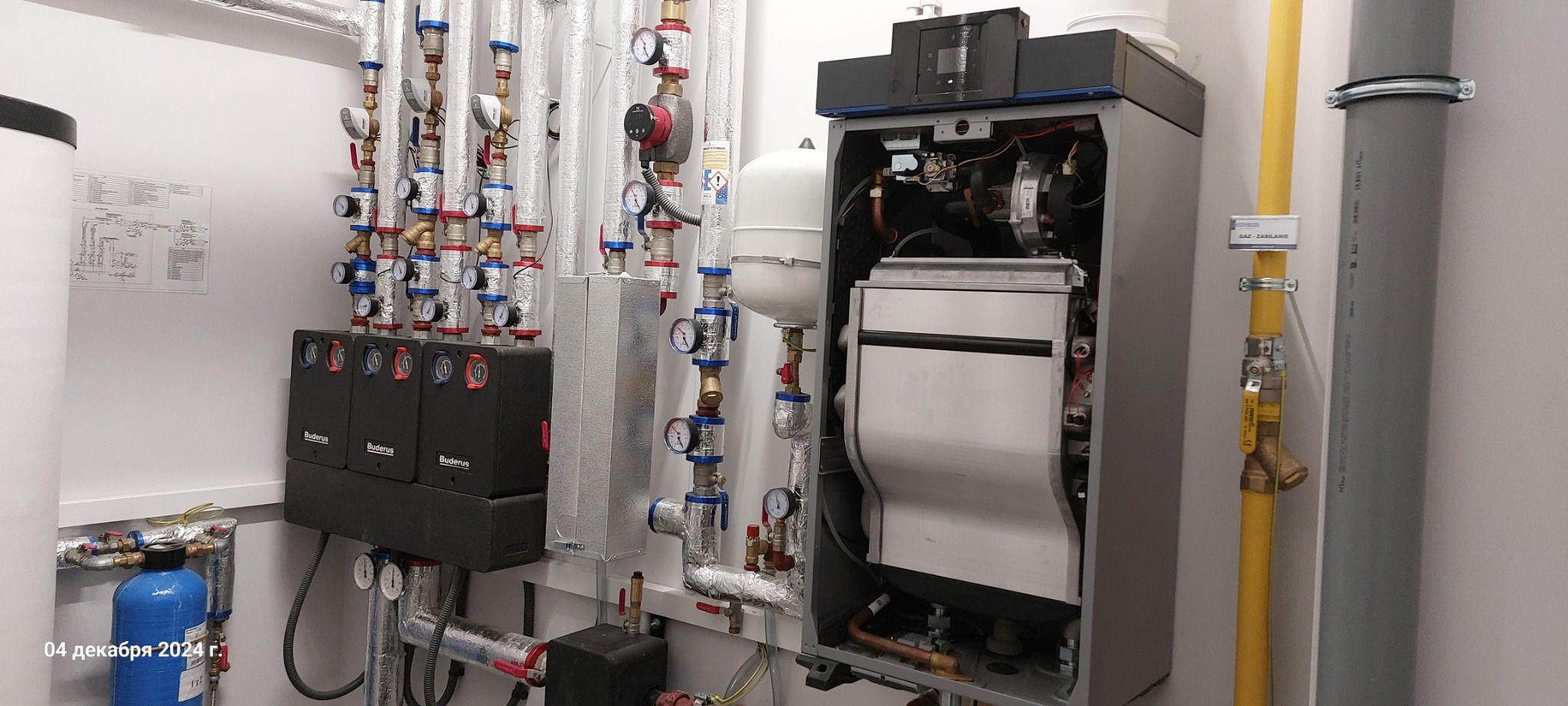 Nowoczesna kotłownia z piecem gazowym, rozdzielaczami Buderus, orurowaniem w izolacji i żółtą rurą gazową z oznaczeniem, widok całości instalacji.