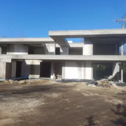 Surowy stan budynku żelbetowego w trakcie budowy, widoczne ściany z bloczków, szalunki stropowe, rusztowanie na dachu, teren budowy z ziemią i paletami.