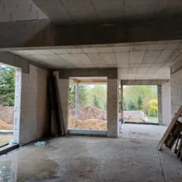 Wnętrze surowego budynku z betonu komórkowego, widoczne przez otwory okienne i drzwiowe hałdy ziemi i drzewa, na podłodze woda, oparte o ścianę drewniane palety.